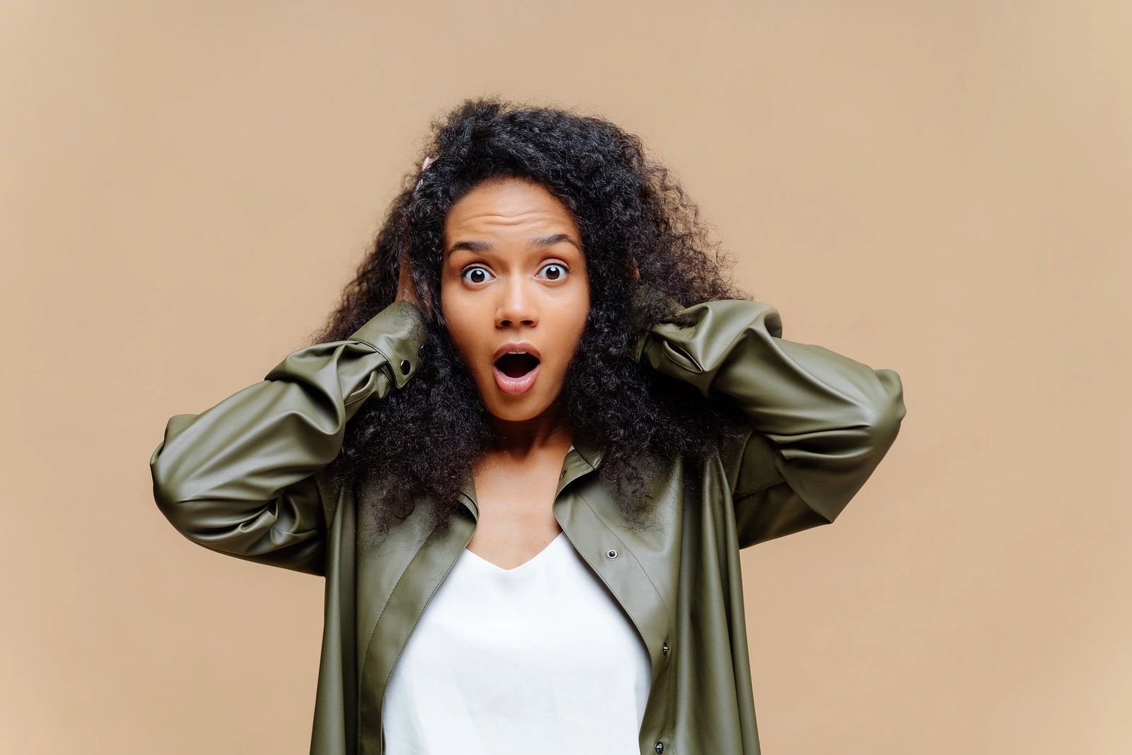 Impressed African American woman keeps both hands on head, opens mouth widely, dressed in white t shirt and leather shirt, being amazed by something, isolated over brown background. Omg concept