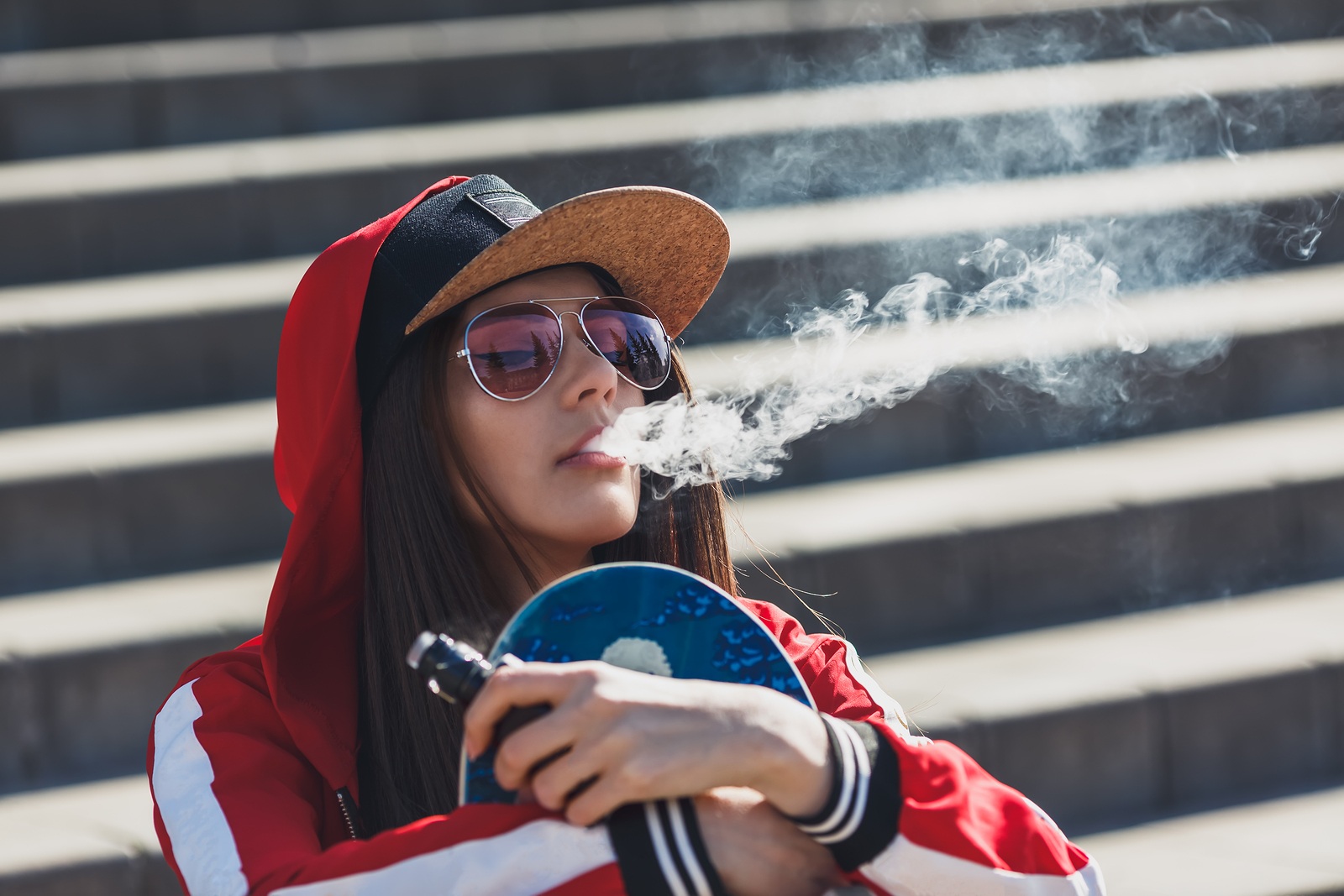 Vaping girl. Young woman with skateboard vape e-cig. Pretty young female in black hat, red clothing vape ecig, vaping device at the sunset. Toned image. Hip hop style.