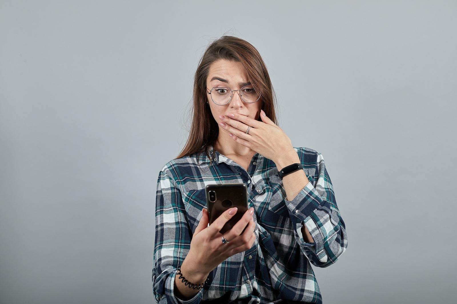 Young brunette girl blue green in checked shirt on gray background, shocked woman with glasses is talking on phone that looks like an iPhone