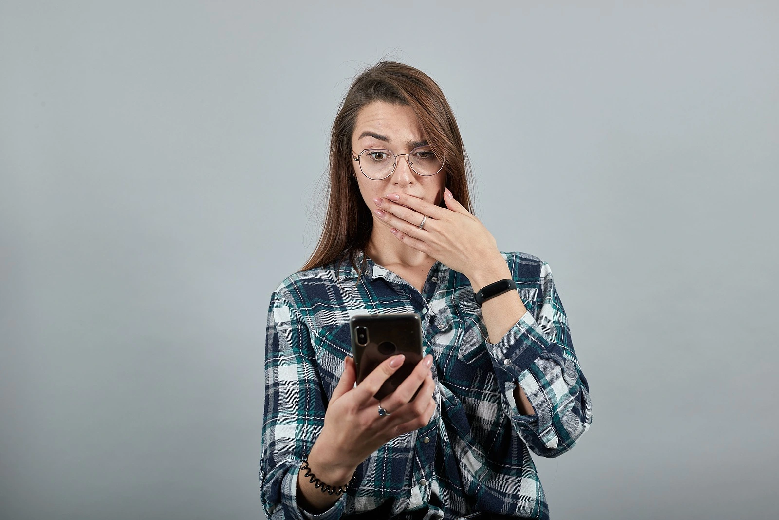 Young brunette girl blue green in checked shirt on gray background, shocked woman with glasses is talking on phone that looks like an iPhone