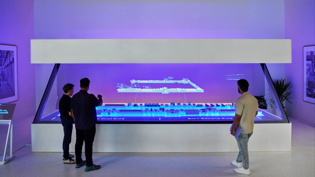 Three people study a holographic display of a production line at Apple's Houston training center