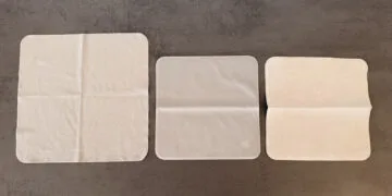 Three Apple polishing cloths side by side on a grey surface, the Apple Vision Pro cloth on the left, the 2026 Apple polishing cloth in the middle and the 2021 version on the right