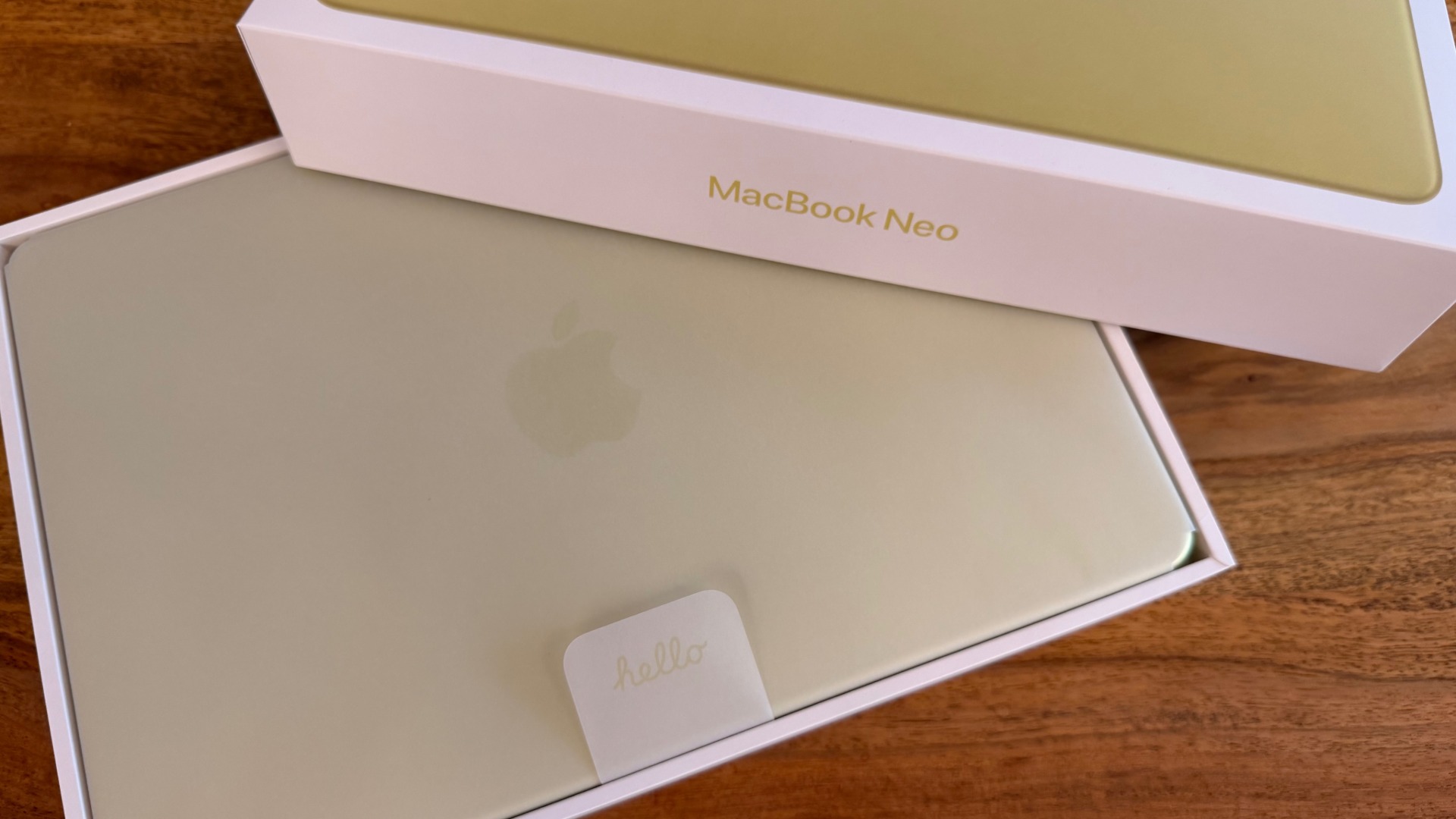 Half-opened box of the MacBook Neo in Citrus, the device still resting in its protective sleeve inside the packaging