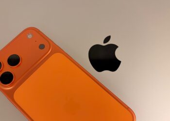 An iPhone 17 Pro Max in Cosmic Orange resting on a silver MacBook Pro next to the Apple logo, the two devices to secure when travelling with an iPhone