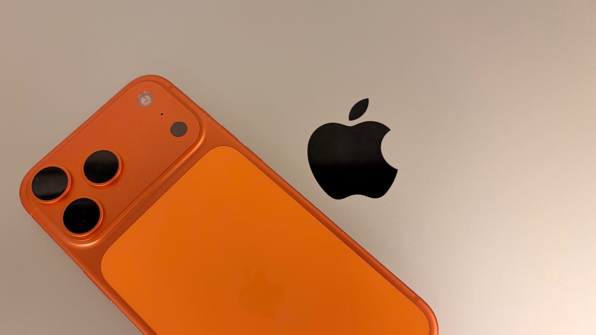 An iPhone 17 Pro Max in Cosmic Orange resting on a silver MacBook Pro next to the Apple logo, the two devices to secure when travelling with an iPhone