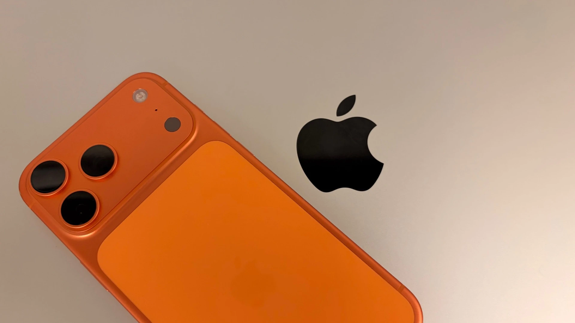 An iPhone 17 Pro Max in Cosmic Orange resting on a silver MacBook Pro next to the Apple logo, the two devices to secure when travelling with an iPhone