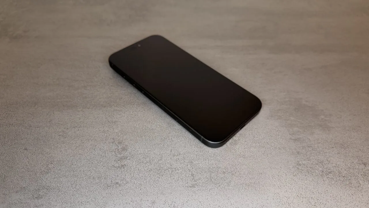 iPhone 20 display comparison: current black iPhone Pro with a flat display, shown at an angle from above on a grey stone surface