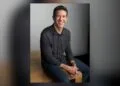 John Ternus sitting on a wooden edge in front of a grey wall, smiling