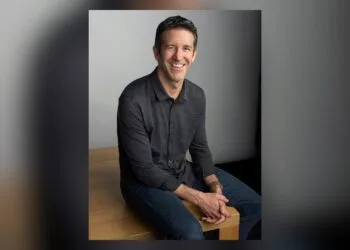 John Ternus sitting on a wooden edge in front of a grey wall, smiling