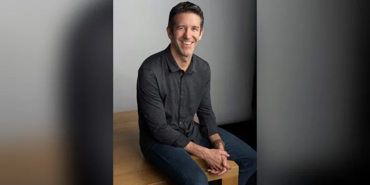 John Ternus sitting on a wooden edge in front of a grey wall, smiling