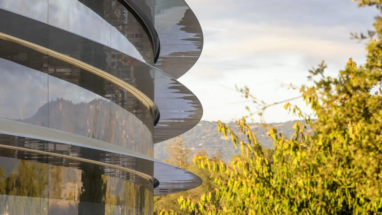 For the Apple DOJ settlement: curved glass facade of Apple Park with cantilevered roof edges, a tree and wooded hills in evening light on the right