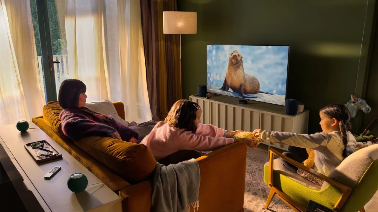 Apple Event Welcome home: a family in a living room watching TV with Apple TV, two HomePod speakers beside the screen and two green HomePod mini speakers behind the sofa