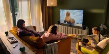 Apple Event Welcome home: a family in a living room watching TV with Apple TV, two HomePod speakers beside the screen and two green HomePod mini speakers behind the sofa