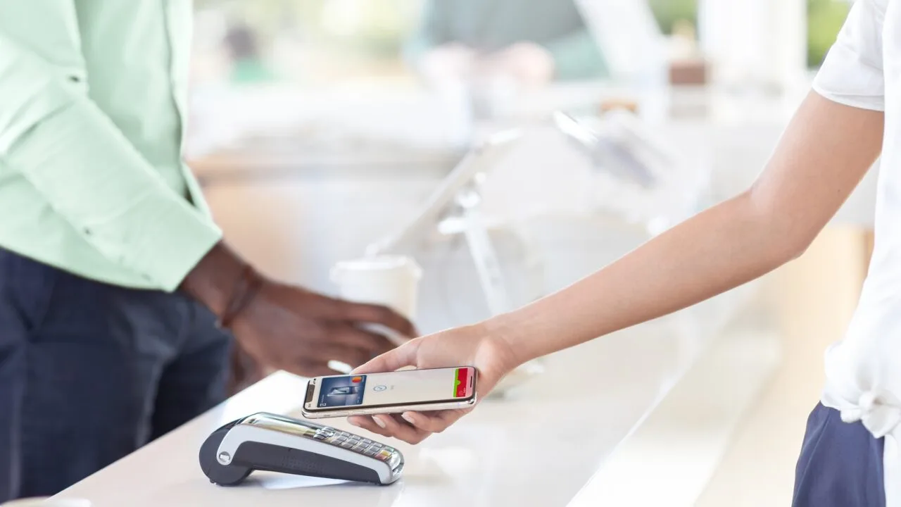 Apple Pay NFC UK: a hand holds an iPhone showing a completed Apple Pay payment over a card terminal on a shop counter