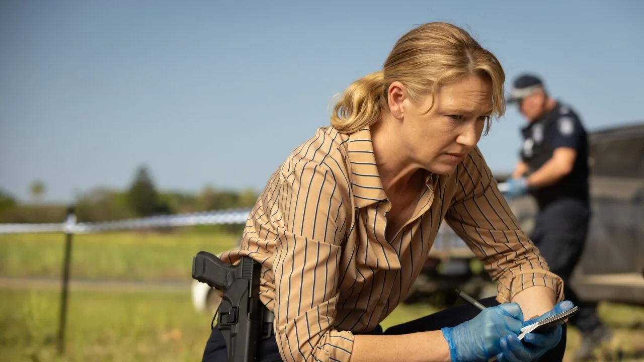 Still from Dustfall on Apple TV: Anna Torv as detective Tig Pollard kneels with a notepad and gloves at a cordoned-off crime scene, a police officer in the background
