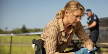 Still from Dustfall on Apple TV: Anna Torv as detective Tig Pollard kneels with a notepad and gloves at a cordoned-off crime scene, a police officer in the background