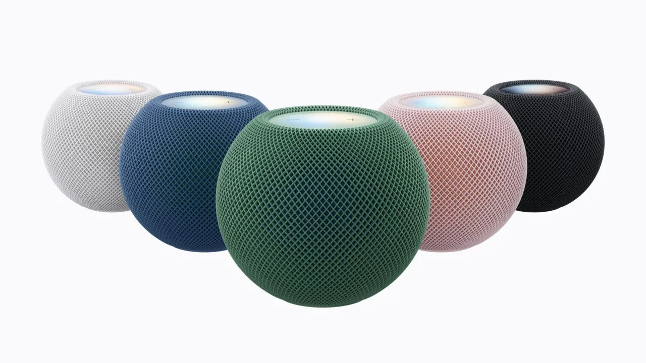 HomePod mini 2 in white, blue, green, pink and midnight arranged in a semicircle, the green model in front with its top lit up