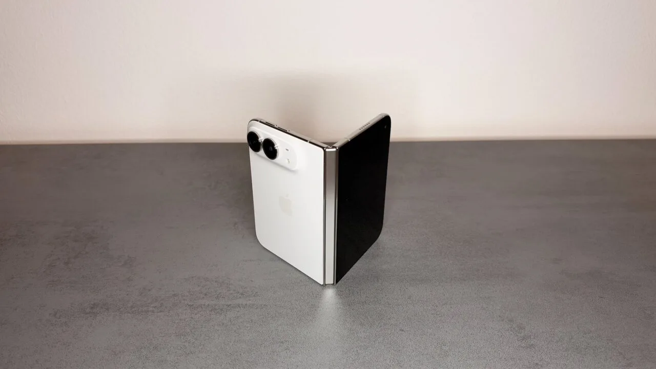 iPhone Duo hinge: a white iPhone Duo half unfolded and standing like a book on a gray stone surface, with the silver hinge facing the camera