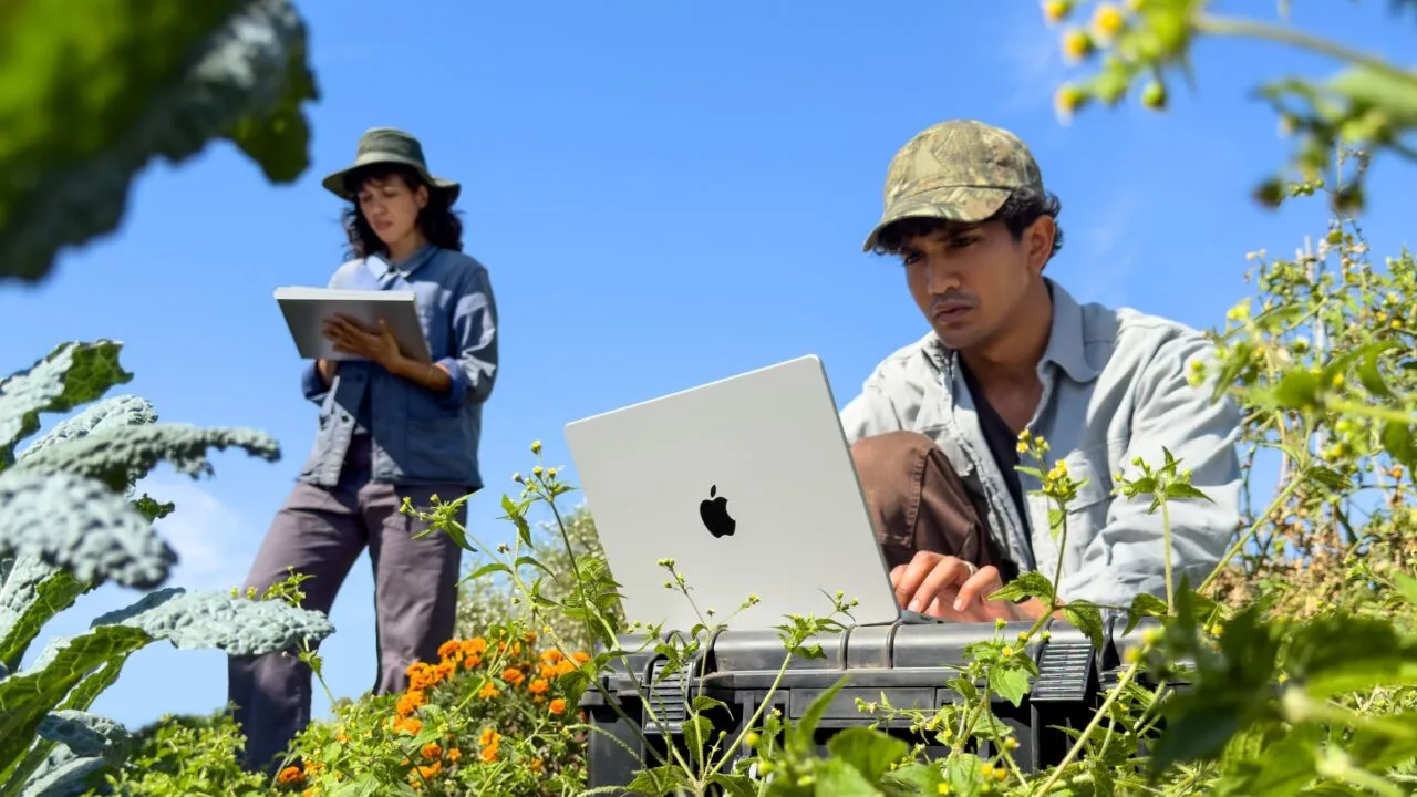 A man works on a silver MacBook Pro M6 among plants while a woman takes notes on an iPad behind him