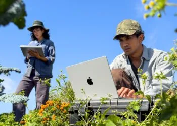A man works on a silver MacBook Pro M6 among plants while a woman takes notes on an iPad behind him