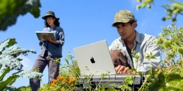 A man works on a silver MacBook Pro M6 among plants while a woman takes notes on an iPad behind him
