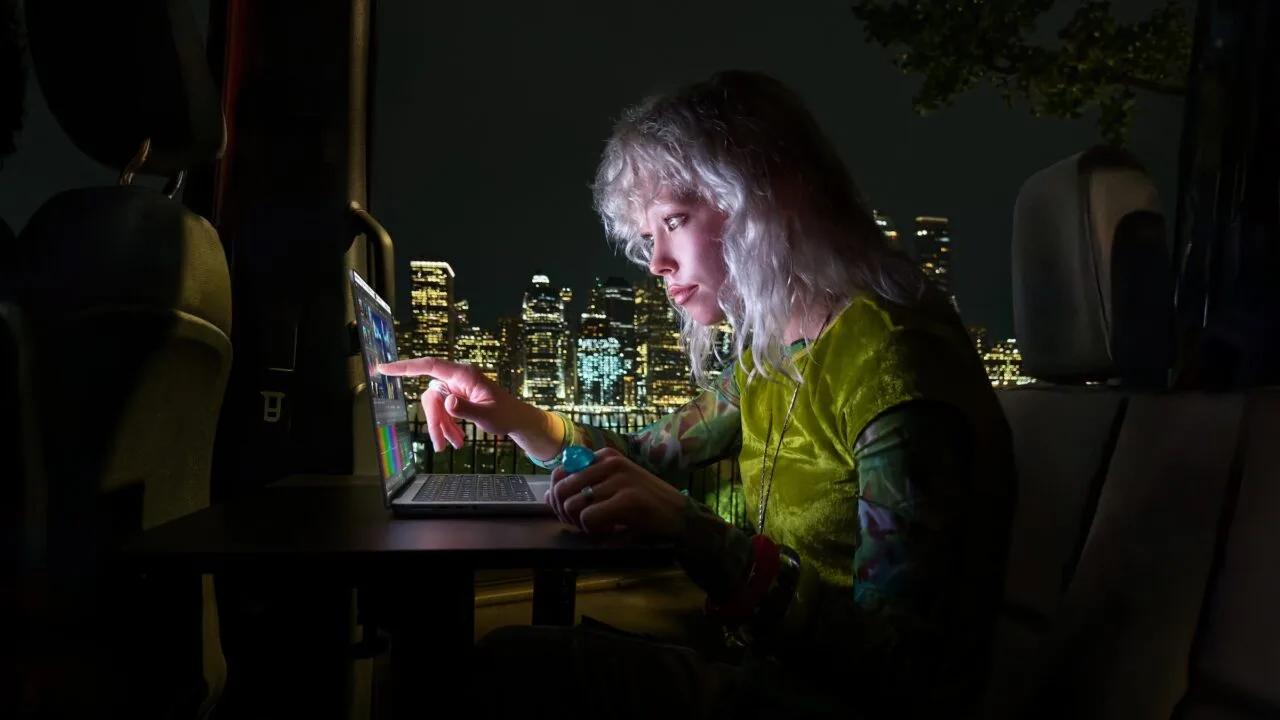 MacBook Pro touch: woman tapping the display of a MacBook Pro with PROLED XDR Display with her finger inside a van at night, a lit-up skyline behind her