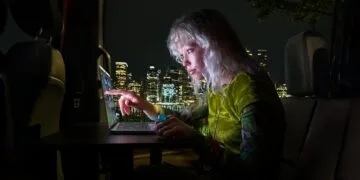 MacBook Pro touch: woman tapping the display of a MacBook Pro with PROLED XDR Display with her finger inside a van at night, a lit-up skyline behind her