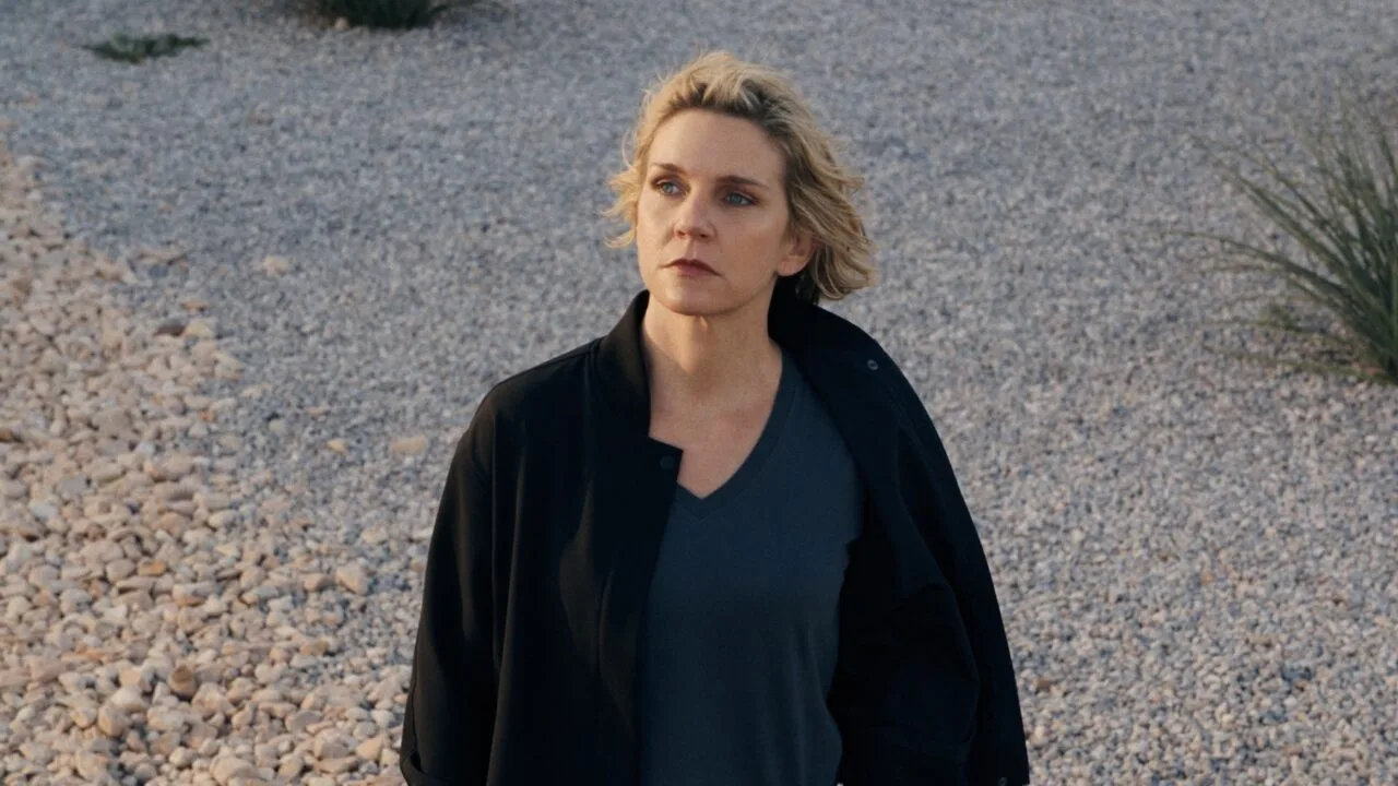 Pluribus season 2: Rhea Seehorn as Carol Sturka in a black jacket standing on a gravel lot with desert plants, scene from the first season