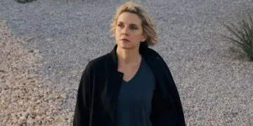 Pluribus season 2: Rhea Seehorn as Carol Sturka in a black jacket standing on a gravel lot with desert plants, scene from the first season