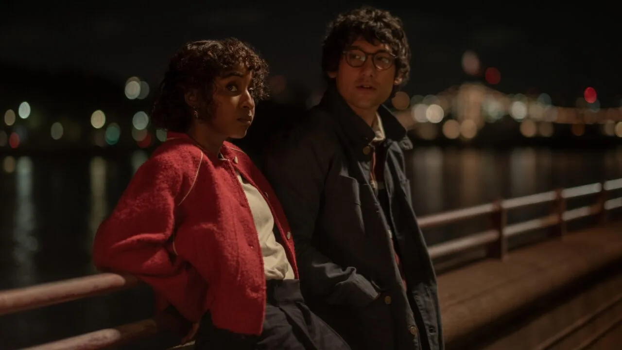 Still from Prodigies: Ayo Edebiri in a red jacket and Will Sharpe in glasses lean on a railing by the water at night, city lights in the background