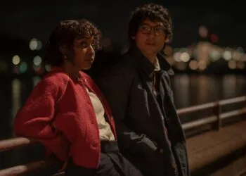 Still from Prodigies: Ayo Edebiri in a red jacket and Will Sharpe in glasses lean on a railing by the water at night, city lights in the background