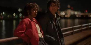 Still from Prodigies: Ayo Edebiri in a red jacket and Will Sharpe in glasses lean on a railing by the water at night, city lights in the background