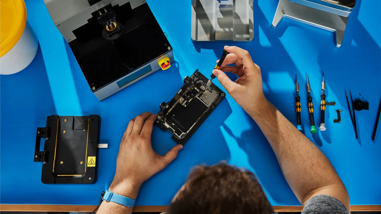 Self Service Repair iPhone 18 Pro: hands repairing an opened iPhone in a holder on a blue workbench, next to Apple repair tools and screwdrivers
