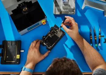 Self Service Repair iPhone 18 Pro: hands repairing an opened iPhone in a holder on a blue workbench, next to Apple repair tools and screwdrivers