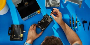 Self Service Repair iPhone 18 Pro: hands repairing an opened iPhone in a holder on a blue workbench, next to Apple repair tools and screwdrivers