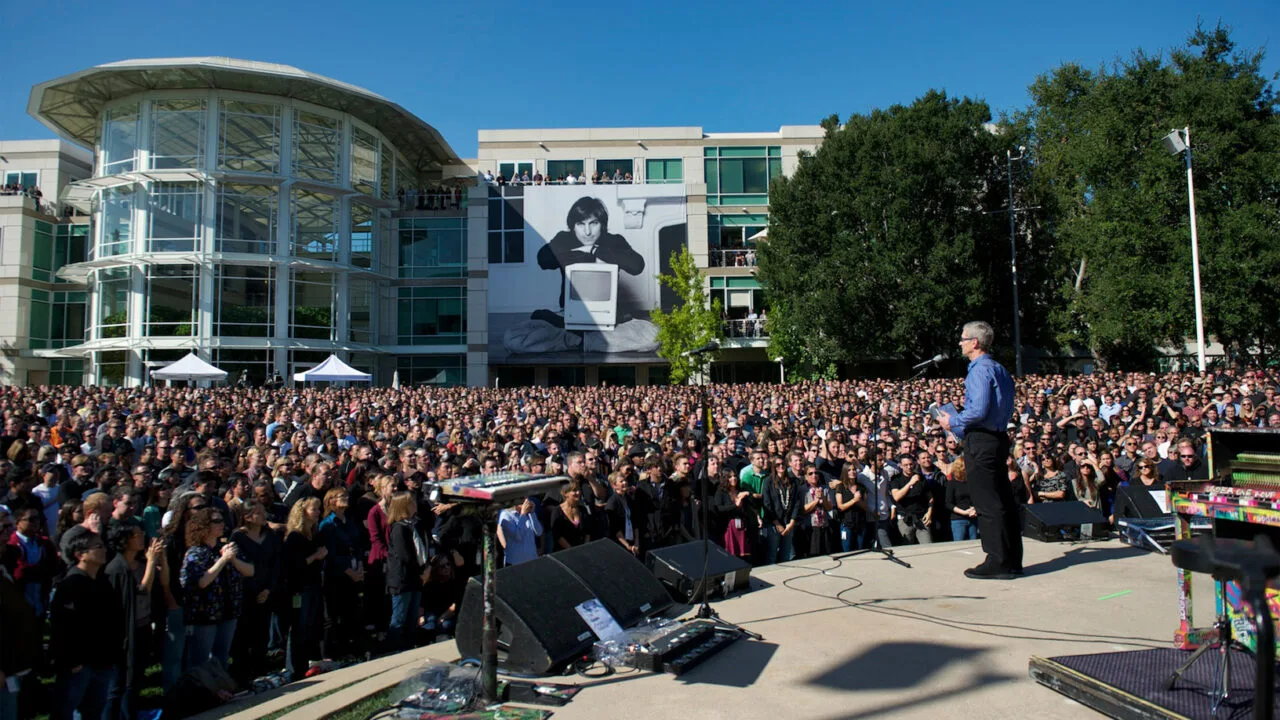 Tim Cook addresses thousands of employees on Apple's campus after Steve Jobs' death in 2011, with a large portrait of Steve Jobs with a Macintosh in the background