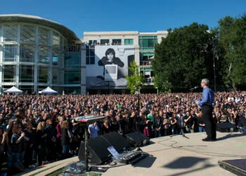 Tim Cook addresses thousands of employees on Apple's campus after Steve Jobs' death in 2011, with a large portrait of Steve Jobs with a Macintosh in the background
