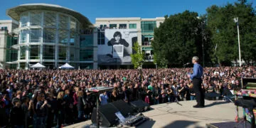 Tim Cook addresses thousands of employees on Apple's campus after Steve Jobs' death in 2011, with a large portrait of Steve Jobs with a Macintosh in the background