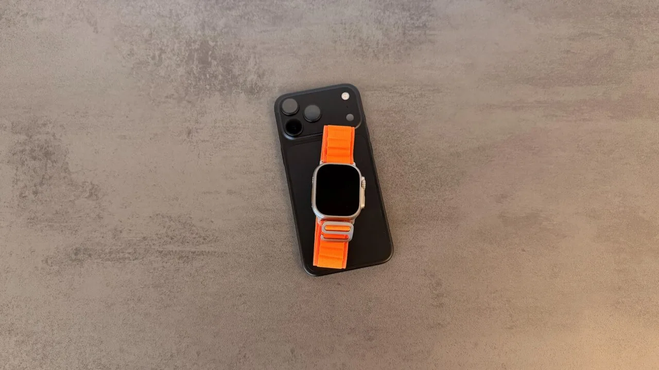 Taptic Engine royalties: black iPhone with camera plateau and an Apple Watch Ultra with an orange Alpine Loop on top, lying on a grey stone table