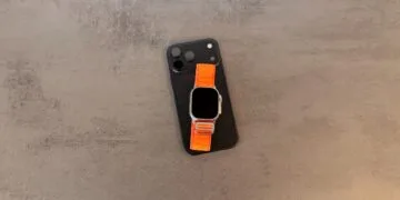 Taptic Engine royalties: black iPhone with camera plateau and an Apple Watch Ultra with an orange Alpine Loop on top, lying on a grey stone table