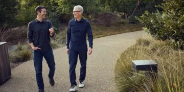 Tim Cook and John Ternus walking side by side and laughing on a path through a green landscape, Ternus on the left, Cook on the right